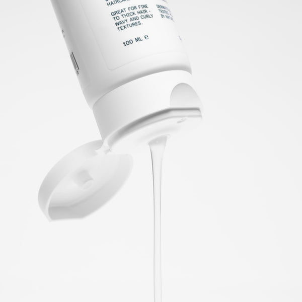 A close-up of a white squeeze bottle of hair care product with its cap off, showing a thin stream of white cream or gel dripping against a clean white background.