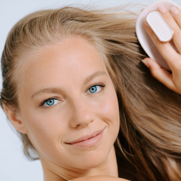 A close-up lifestyle shot of a woman with light brown hair using a white hairbrush to brush her hair, set against a clean light gray background.