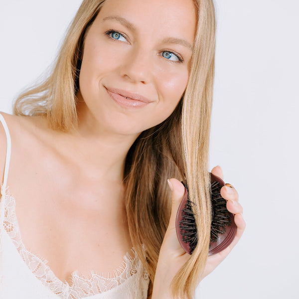 A blonde woman with long, straight hair holds a dark brown hairbrush against her hair, wearing a white lace-trim camisole against a plain white background.