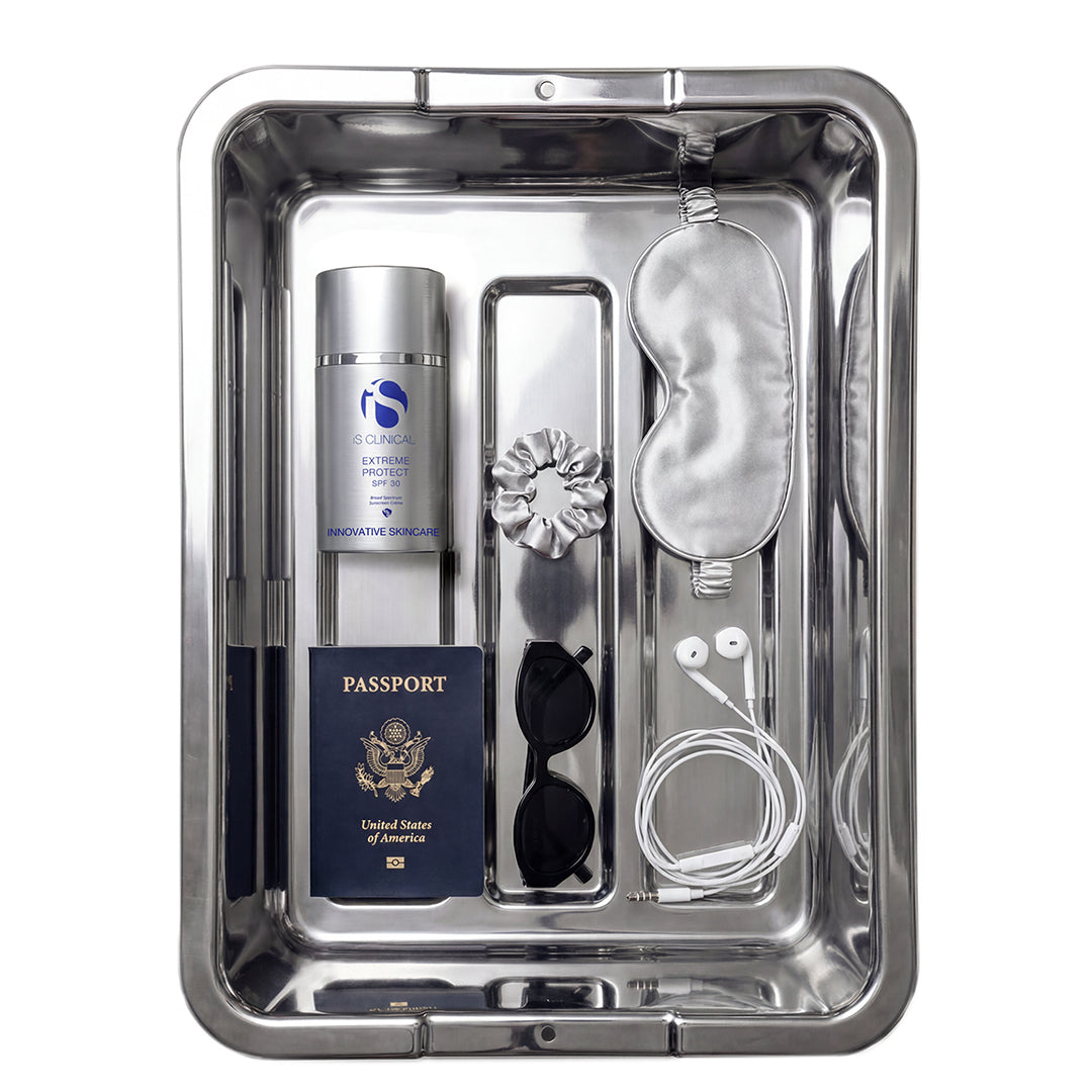 A stainless steel travel organizer tray containing a silver travel spray bottle, a navy blue United States passport, black sunglasses, white wired earbuds, a silver sleep mask, a silver flower-shaped hair clip, and a white cord, all arranged neatly on a white background.