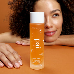 Woman with curly hair leaning on an orange surface with a Pai skincare product bottle in front.