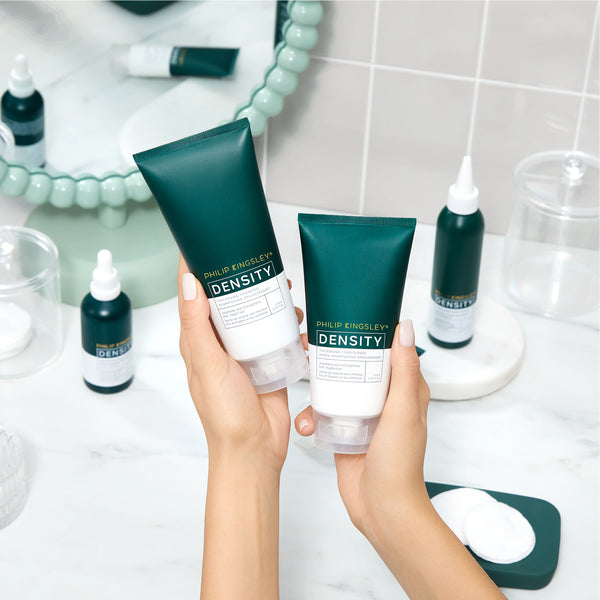 Hands holding Philip Kingsley Density hair care products with a bathroom setting in the background.
