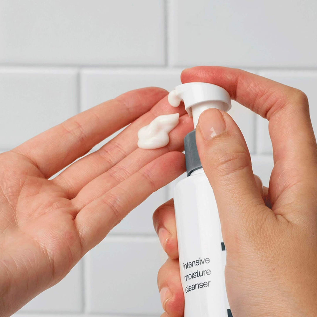 Person applying cream from a bottle with a pump against a white tiled wall.