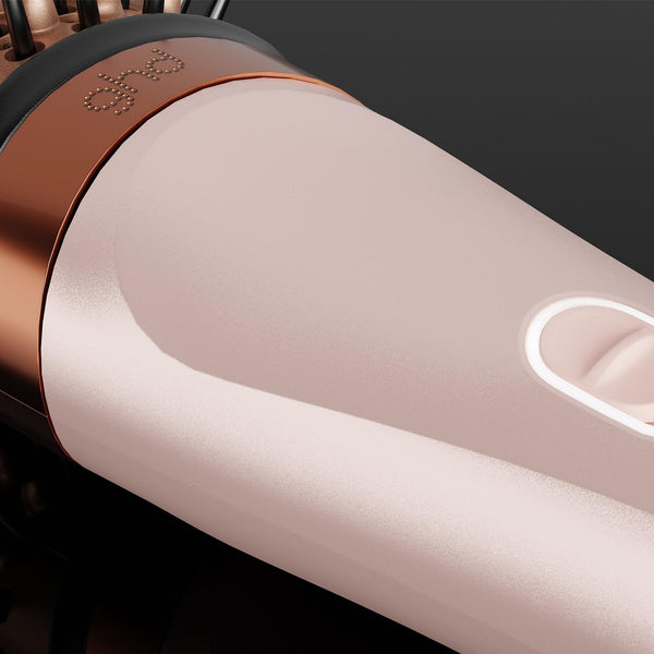 A close-up of a GHD hair styling tool, showing its metallic rose gold barrel, a glowing white power switch, and a copper-colored trim against a dark background.