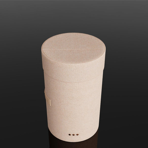 A minimalist, cylindrical container or speaker in a light beige fabric texture, standing upright on a reflective black surface against a dark gradient background.