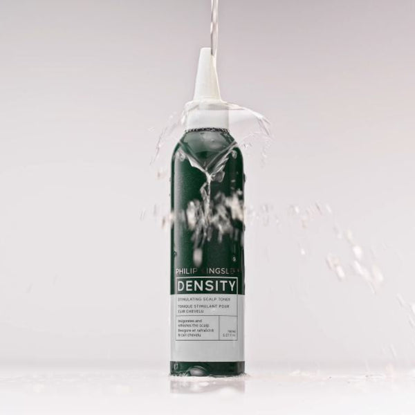 A single cylindrical bottle of DENSITY brand product stands upright on a white surface against a light gray studio background, with water splashing around it and a clear stream entering the top opening.