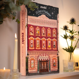 A hardcover book titled 'zooki 28 Days of Radiance' stands upright on a shelf, surrounded by candles and a glass vase with flowers in a warm, styled interior setting.