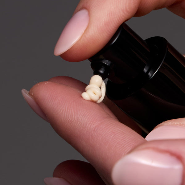 A close-up of a hand with pale pink manicured nails dispensing a small dollop of cream-colored cosmetic product from a black pump bottle against a dark gray background.