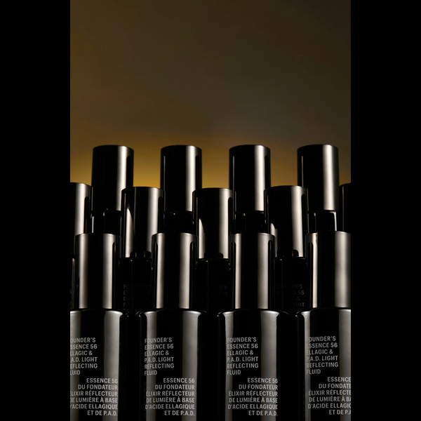 A group of black cosmetic bottles with silver caps arranged in a row against a dark background with a warm golden glow near the top. The bottles have white text on the lower section that reads 'BUNDER'S ESSENCE 56 LUMIÈRE & LUMIÈRE REFLECTING'.