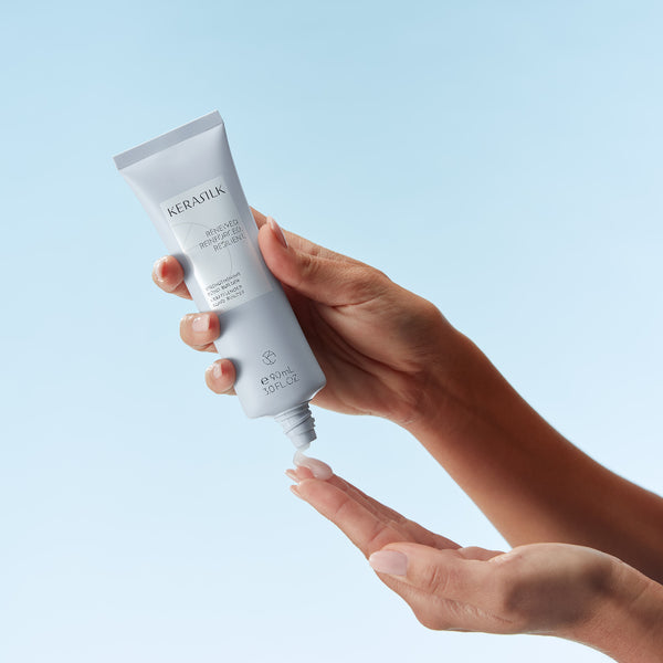 Hand holding a tube of KeraSilk hair care product against a light blue background