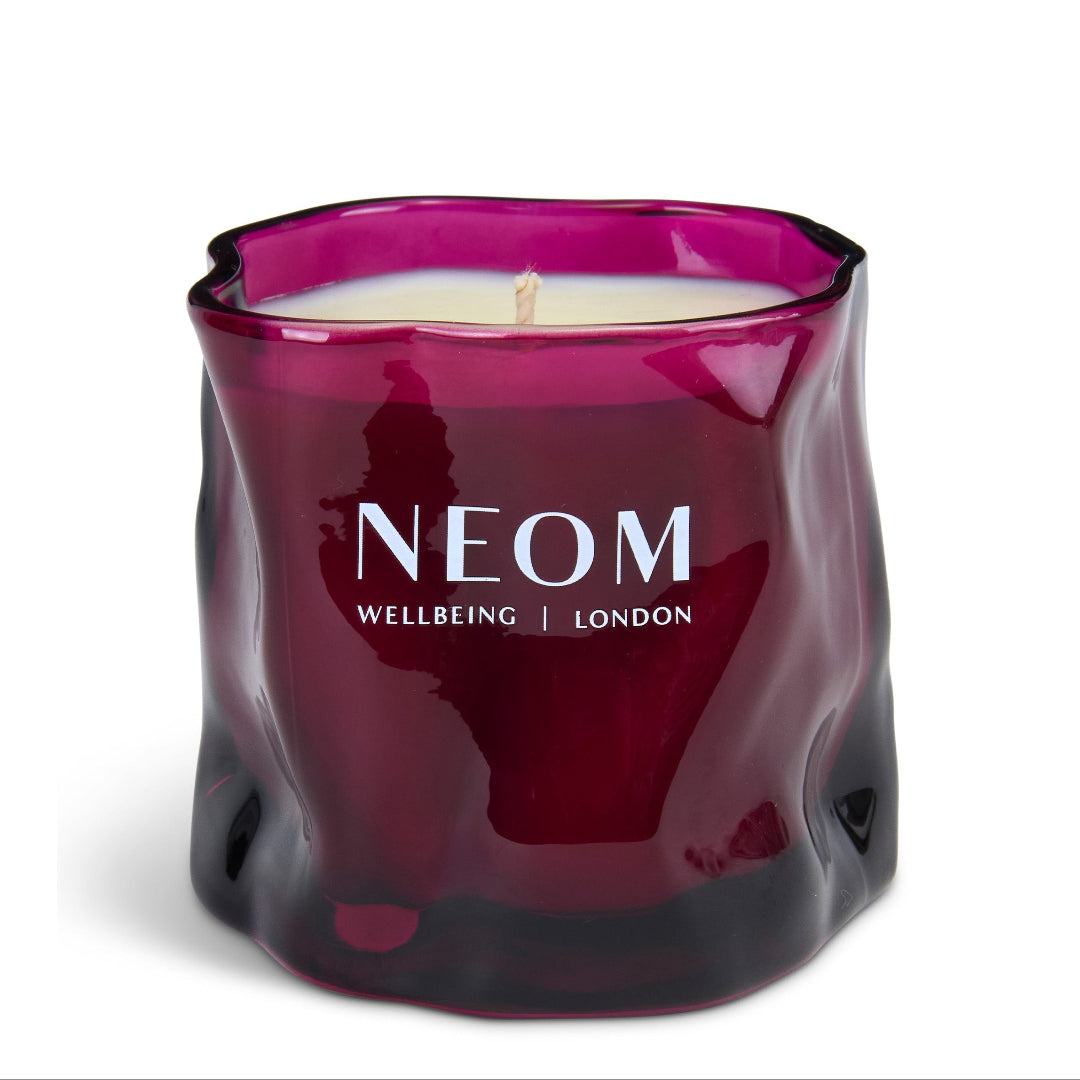 A NEOM Wellbeing London scented candle in a wavy, wavy textured dark magenta glass jar with white wax and a single wick, on a clean white background.