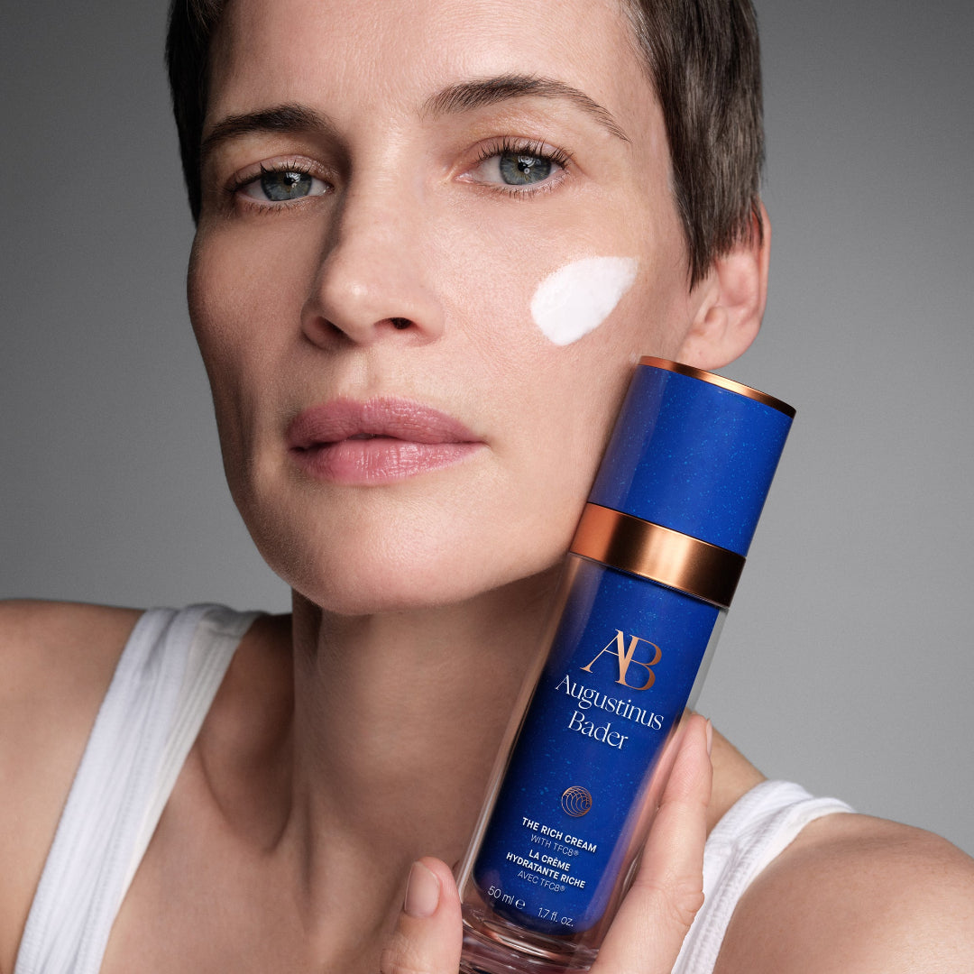 A close-up of a woman holding a blue and gold bottle of Augustinus Bicker The Rich Cream against a neutral gray background.