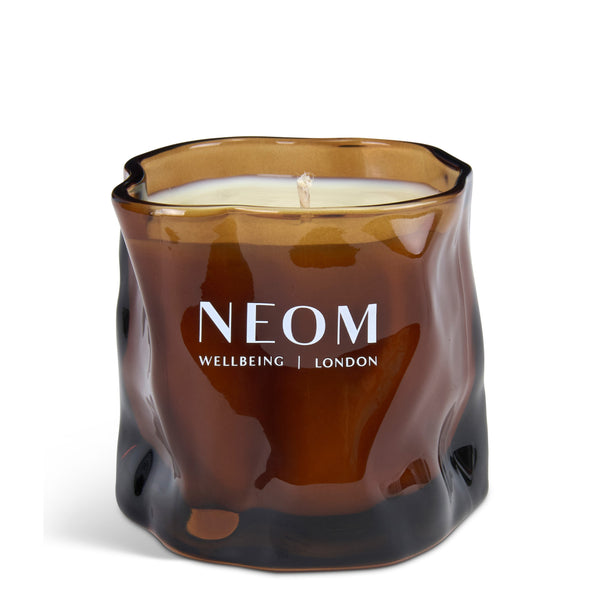 A NEOM candle in a sculptural, wavy amber glass jar with white wax and white printed branding, set against a plain white background.