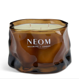 A NEOM Wellbeing London scented candle in an organic, wavy amber-brown glass container, featuring a cream-colored wax fill and white brand lettering on the front.