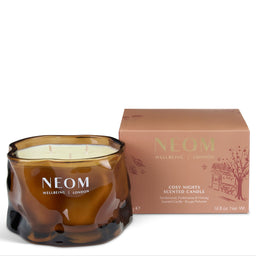 A NEOM Wellbeing London scented candle in a textured amber glass jar, shown next to its dusty rose and gold packaging box on a white background.