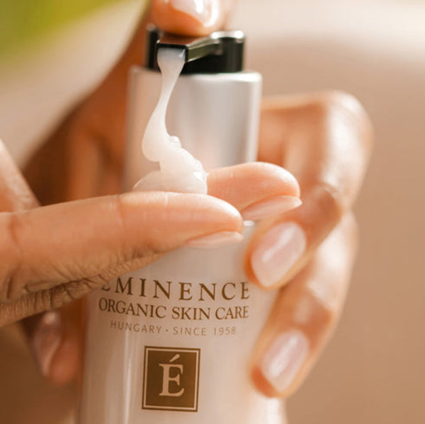 A close-up of hands dispensing white cream from a silver bottle labeled 'MINIMENCE ORGANIC SKIN CARE' against a soft beige background.
