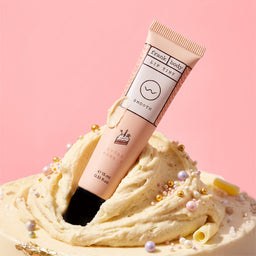 A tube of Frank body Lip Stink SUGAR HAZE Smooth lip tint, standing upright in a swirl of beige cream with decorative pearls and curls against a solid pink background.