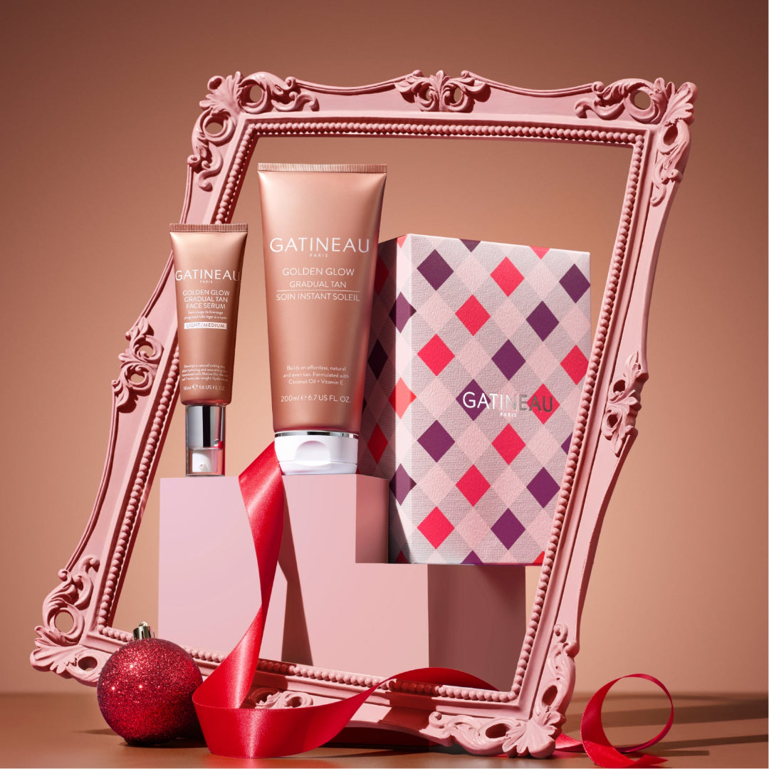 Gatineau Golden Glow Volumizing Hair Product set displayed in an ornate pink frame, featuring a volumizing mousse tube, a matching hair conditioner bottle, and a patterned gift box with a red ribbon and a red ornament on a warm beige background.
