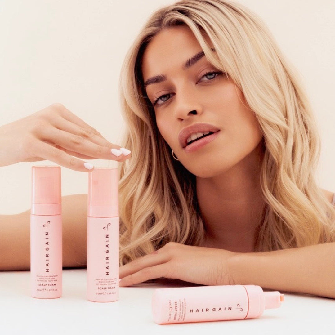 Woman with blonde hair holding two pink bottles of Hairgain product on a white background