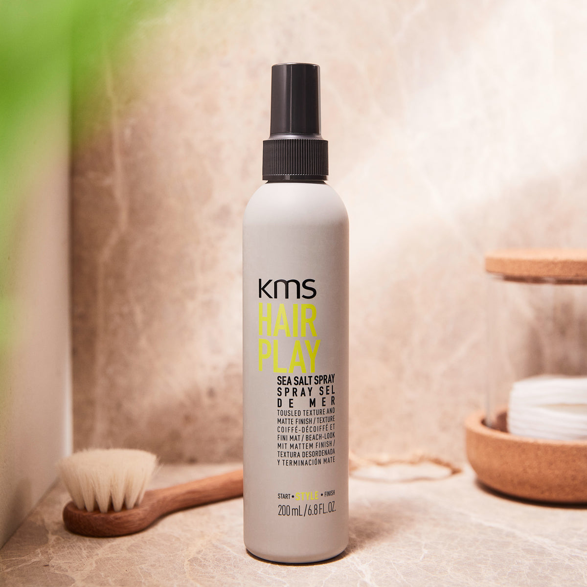 KMS Hair Play Sea Salt Spray bottle on a bathroom counter with a brush and soap dish in the background.