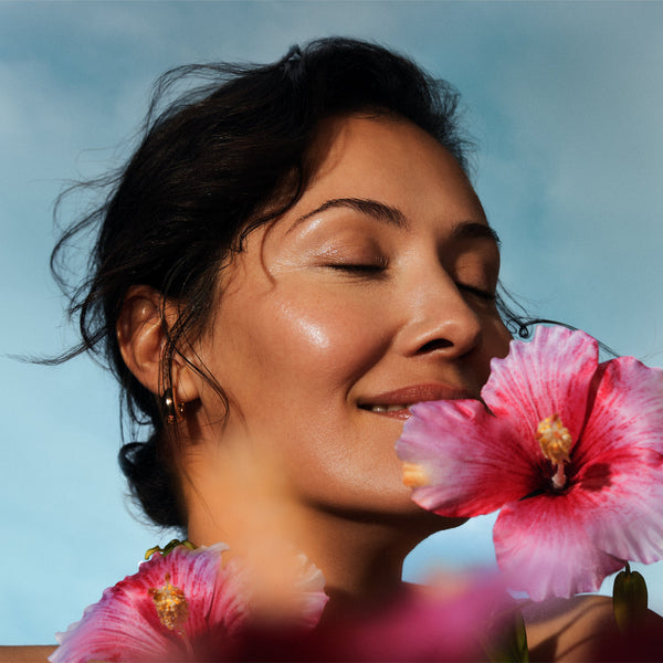 Woman with eyes closed, smelling a pink flower against a clear blue sky