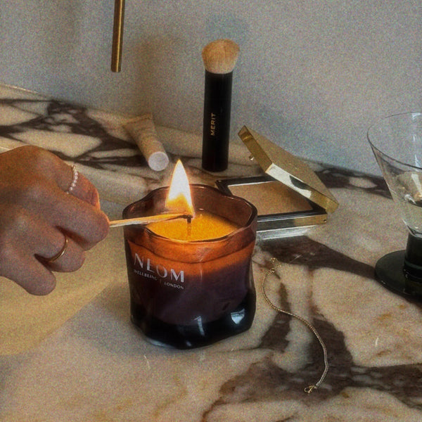 A hand lights a candle in a dark amber glass jar with a visible flame. The candle jar has the brand name 'ALOM' printed on the front.