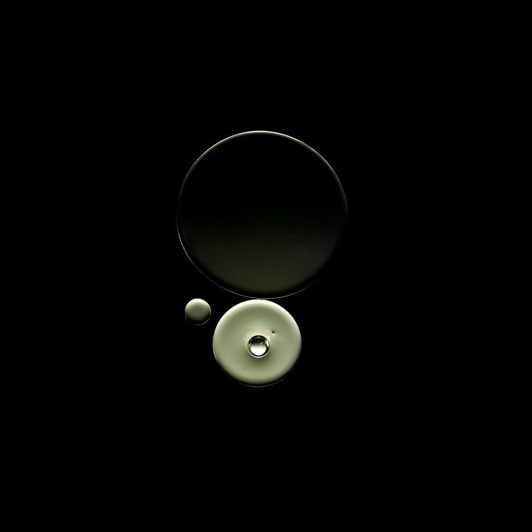 Three circular glass objects, including two larger dark-toned pieces and one pale greenish-beige piece with a small clear droplet, arranged on a solid black background.