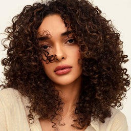 A close-up portrait of a woman with voluminous, tightly curled dark brown hair, wearing a light cream-colored top against a plain off-white background.