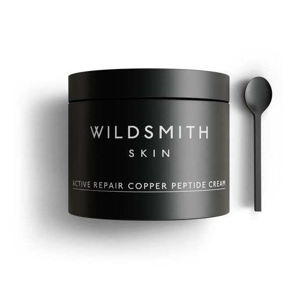 A matte black cosmetic jar labeled 'WILDSMITH SKIN' with white text reading 'ACTIVE REPAIR COPPER PEPTIDE CREAM,' shown on a white background with a matching black spoon to the right.