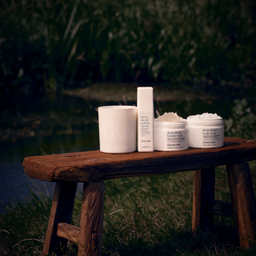 Four skincare products from deep-drip beauty by Dr. Lipp are arranged on a rustic wooden bench outdoors near grass and water.