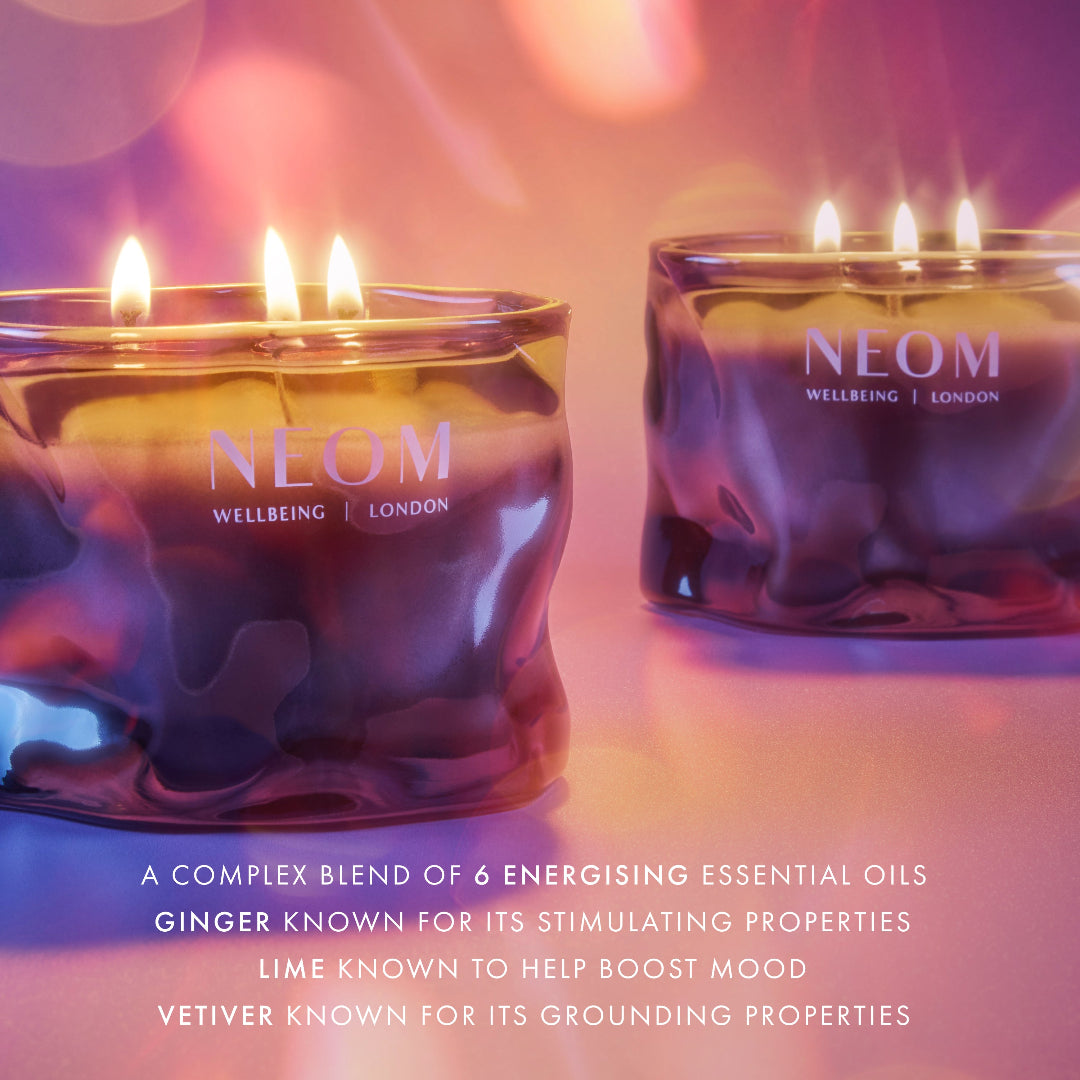 Two lit NEOM Wellbeing London candles in textured amber glass jars, with warm pink and purple lighting and text at the bottom describing the essential oil blend.
