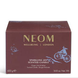 A rectangular NEOM Wellbeing London scented candle box in a deep burgundy-brown color, featuring gold lettering and light blue line-art illustrations of a cyclist and botanical motifs, set against a plain white background.