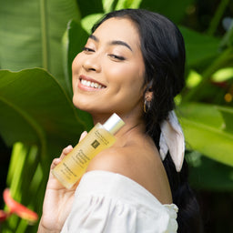 A woman outdoors in front of large green leaves holds a bottle of Essence of Paris Rosemary Hair Growth Treatment. She is smiling and looking upward, wearing a white off-the-shoulder top and gold hoop earrings.