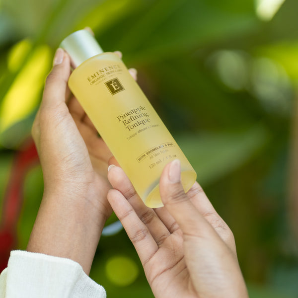 A person holds a bottle of Eminence Haircare Pineapple Defining Tonic against a soft, blurred green leafy background. The product is a translucent yellow liquid in a frosted bottle with a white cap.