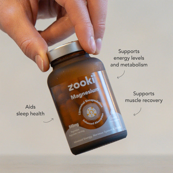 A hand holds a brown amber bottle of Zooki Magnesium supplement. The bottle has a silver cap and a label with white and gold text.