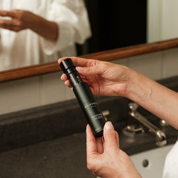 Person holding a black skincare bottle labeled 'Alvera' in front of a mirror.