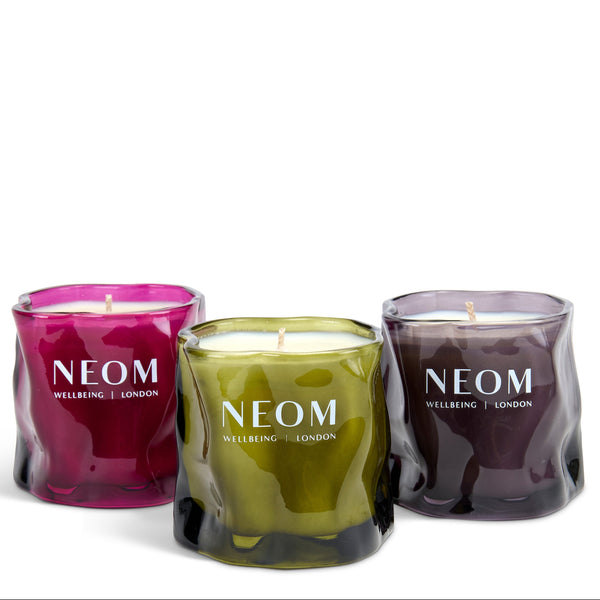 Three wavy glass jar candles in magenta, olive green, and dark plum purple, arranged together on a white background. Each candle features the white printed brand name 'NEOM WELLBEING | LONDON'.