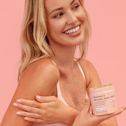 A smiling blonde woman in a white tank top holds a jar of Fresh Body Excessive Whipped Body Scrub, with scrub visible on her skin and the jar against a solid pink background.