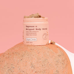A pink tub of Express-o Whipped Body Scrub by frank body sits on top of a mound of textured body scrub against a solid pink background.