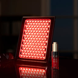 A red LED light therapy panel glowing brightly next to a red and gold skincare bottle on a reflective dark surface.