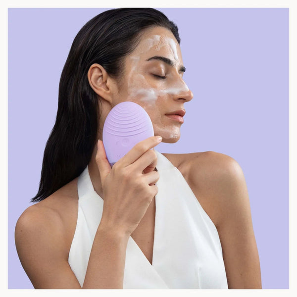A woman with white face cream on her cheek holds a lavender purple silicone facial cleansing brush against her face, set against a solid light purple background.