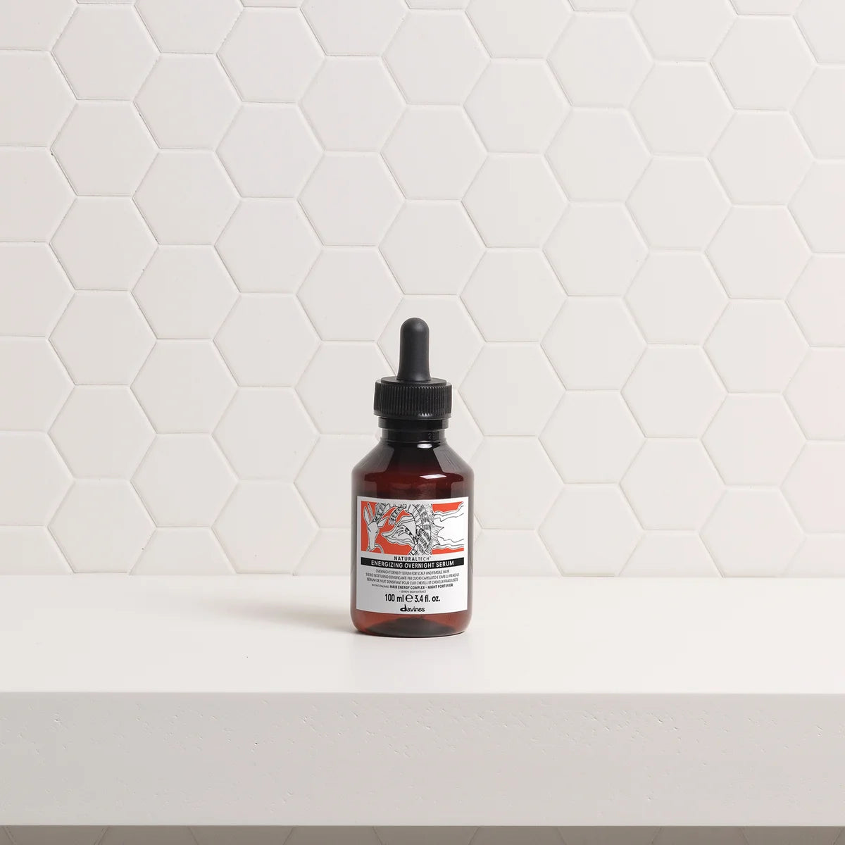 A small amber glass dropper bottle with a black cap stands on a white shelf against a white hexagonal tile wall. The bottle has a white label with red and black text and graphics.