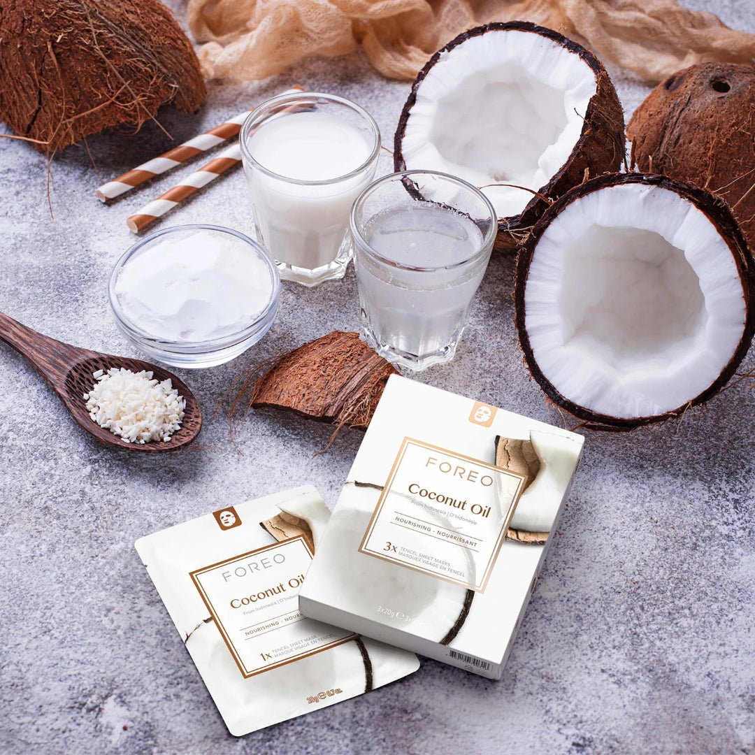 FOREO Coconut Oil skincare packaging shown with whole and halved coconuts, a small bowl of coconut cream, two glasses of white coconut liquid, and a wooden spoon of shredded coconut on a textured gray surface.