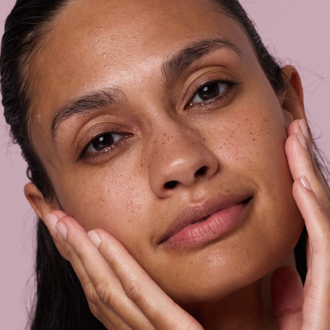 Close-up of a woman's face with hands gently touching her skin against a pink background