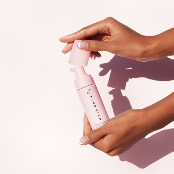 Hand holding a pink bottle with 'Hair Gain' branding against a light background